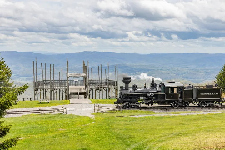 Steam train on tracks near wooden structure with mountains in the background.