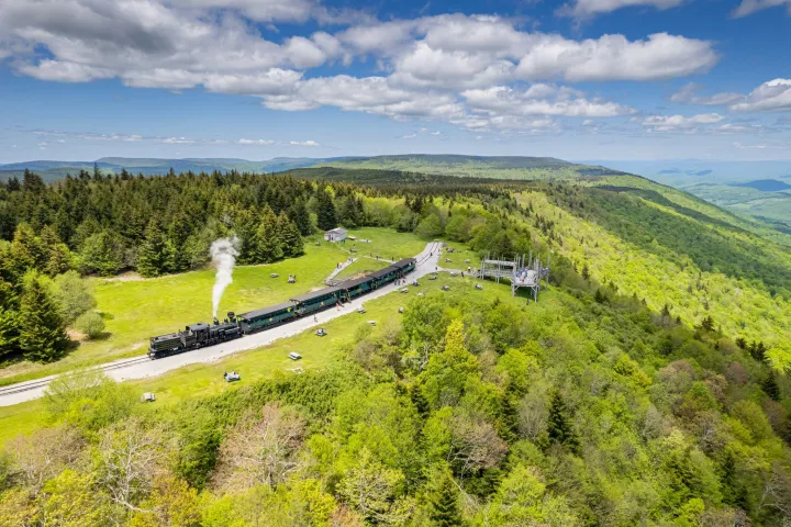 Aerial view of a steam train on tracks through a lush green forested mountain landscape under a cloudy sky.