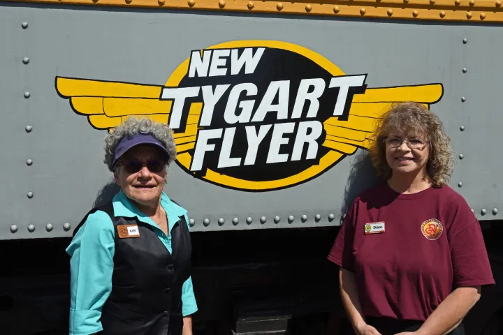 Car hosts Mary Richards and Diane Cash standing in front of the New Tygart Flyer sign.