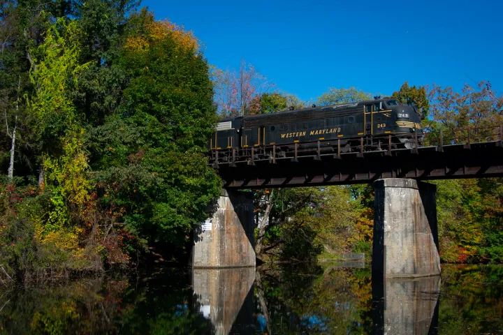 Train on bridge over river with trees and clear blue sky.
