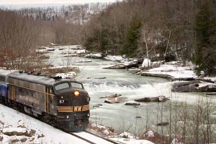 Train traveling near a snowy riverbank in a forested area during winter.