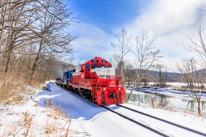 Red train traveling on snowy tracks alongside a river on a clear winter day.