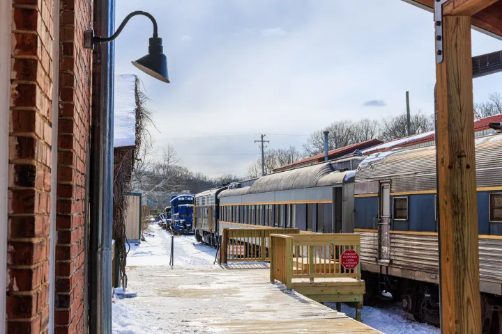Snow-covered train station platform with blue train in background, bordered by brick building.