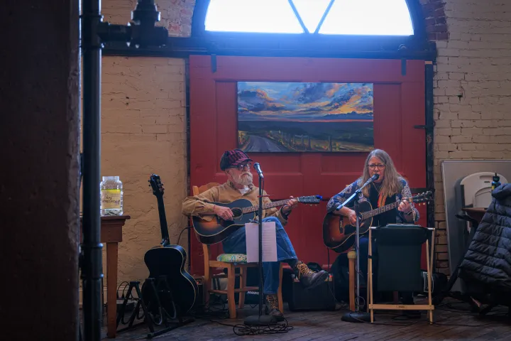 Two musicians playing guitars indoors with a red door and painting in the background.