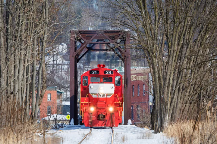 Red train number 24 on snowy tracks, framed by leafless trees and an old building in the background.