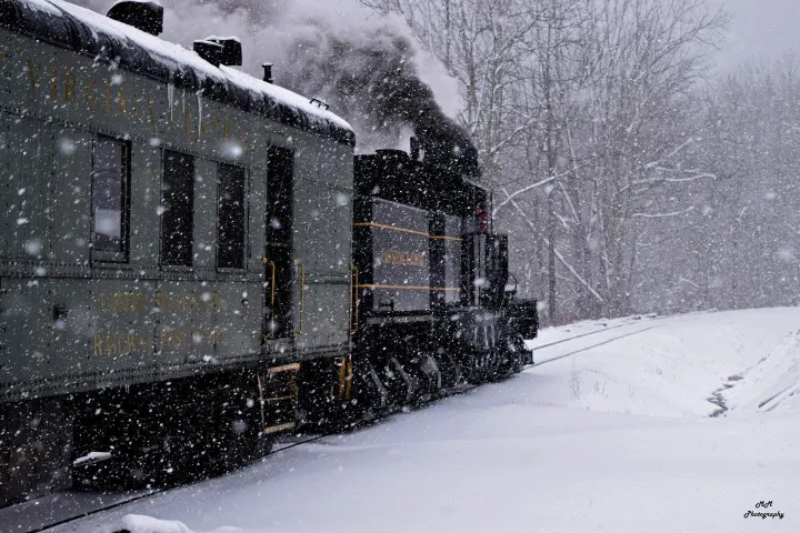 Steam train moving through snowy landscape with trees in the background.