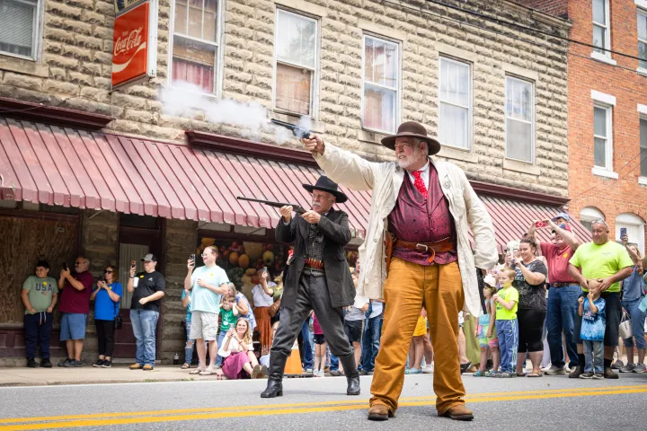 Two actors in Western costumes fire guns in front of a large crowd during a street performance.
