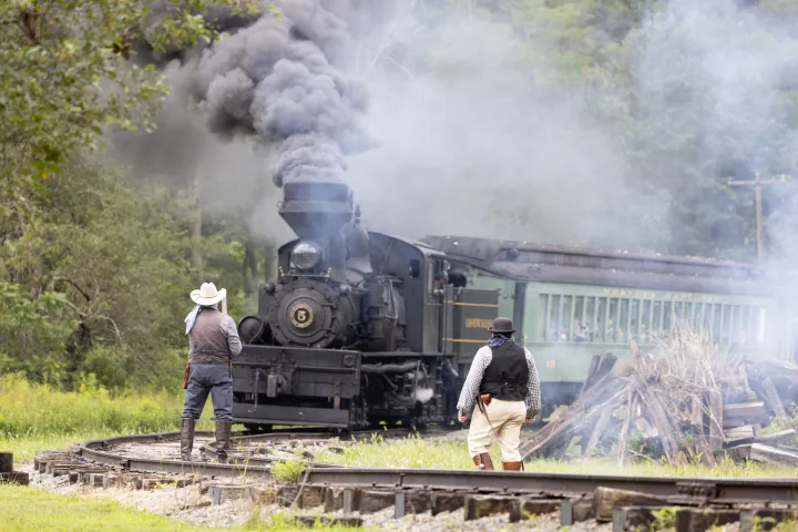 Two people in vintage attire watch a steam train emitting smoke on a curved track.