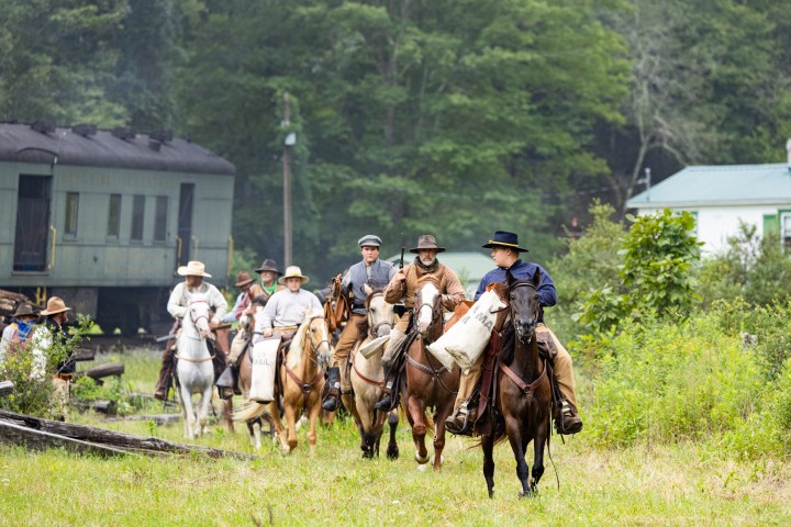 Group of people on horseback with mail bags near a train and greenery.