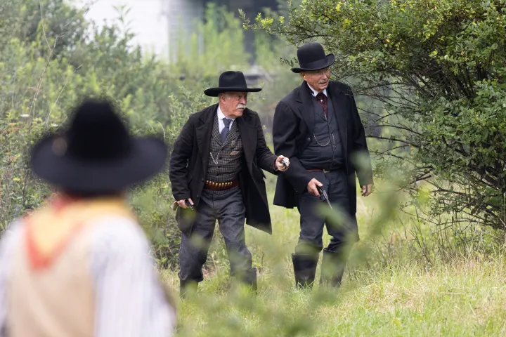 Two men in vintage Western outfits with guns, walking through greenery.
