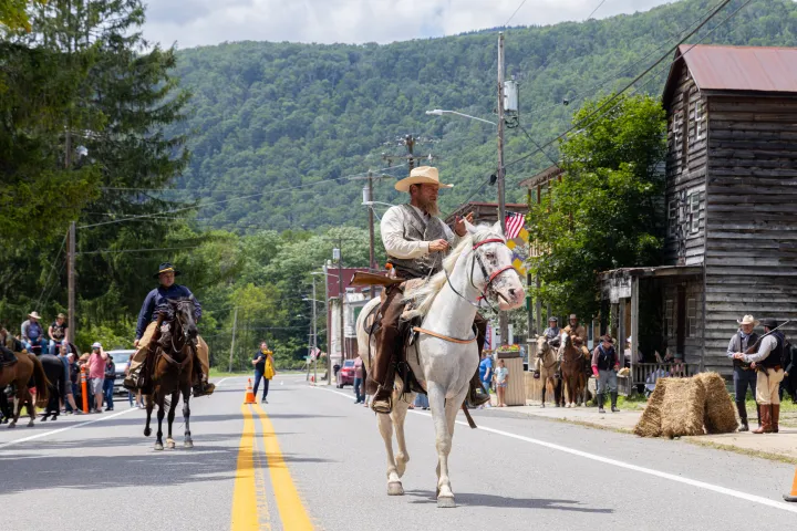 Men in cowboy attire riding horses on a street in a rural town with mountains.