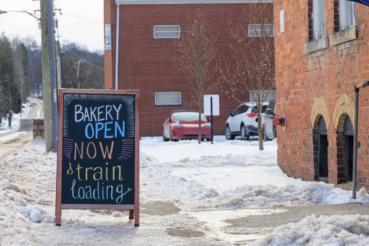 Chalkboard on snowy sidewalk reads 'Bakery Open Now, $train Loading' near brick building.