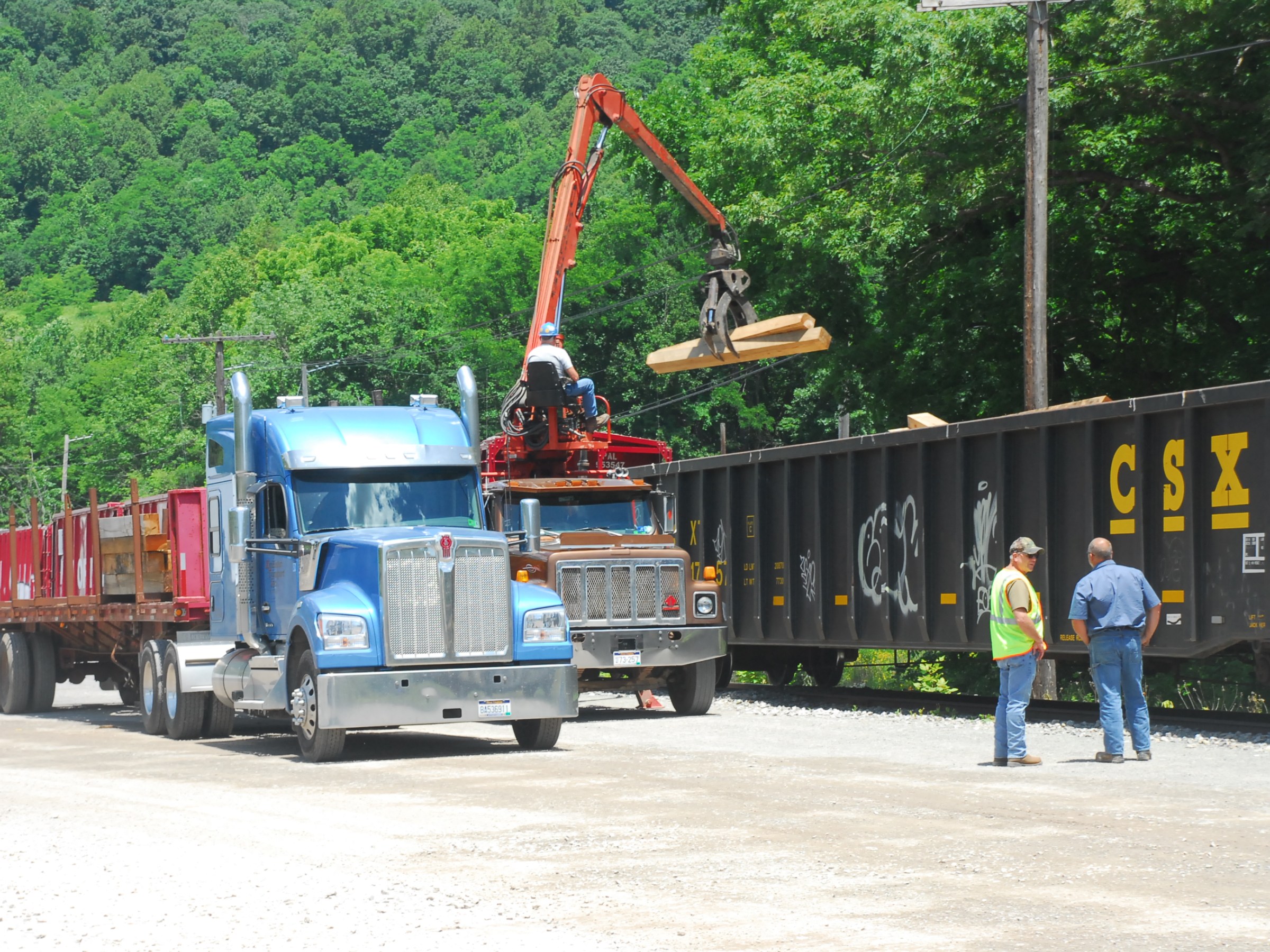 Truck loading equipment near train car with workers and wooded hill in background.
