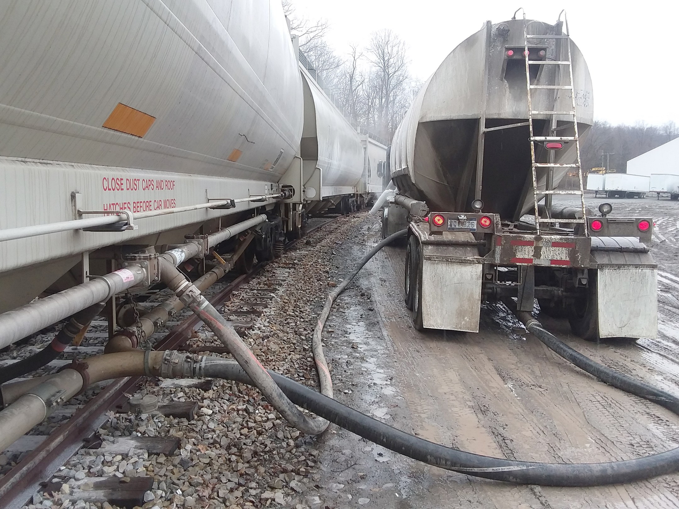 Tanker truck and train connected by hoses on muddy ground.