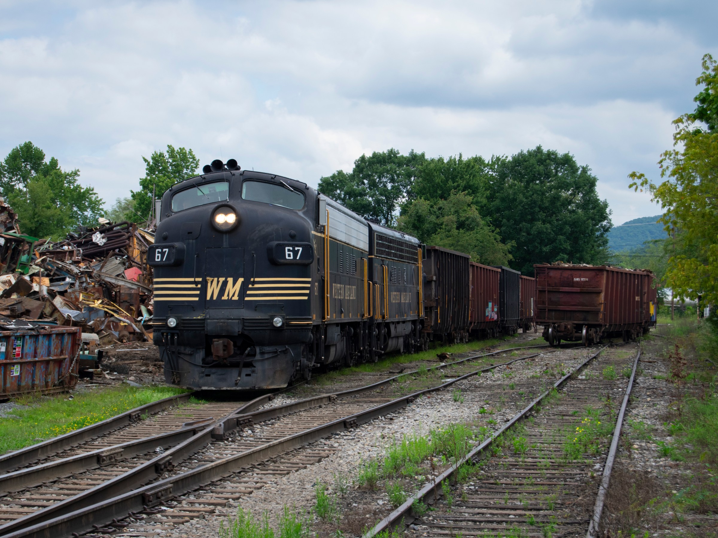 Vintage train on tracks with scrap metal and trees in the background.