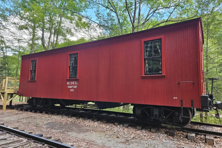 Red wooden train car labeled 'M.L.BR Co. Camp Car 419' on rails in forest setting.
