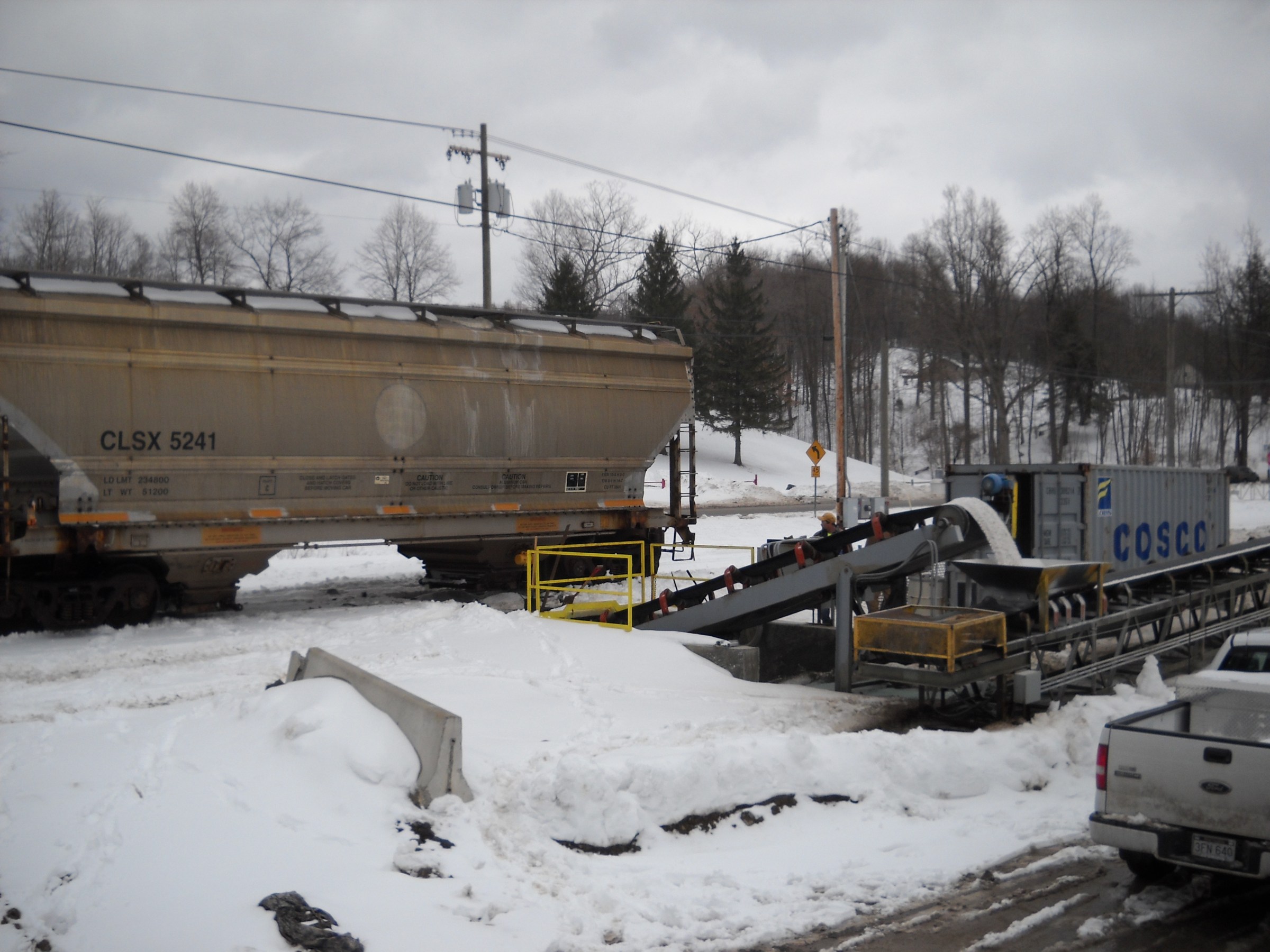 Train car beside snow-covered conveyor system unloading salt under a cloudy sky.