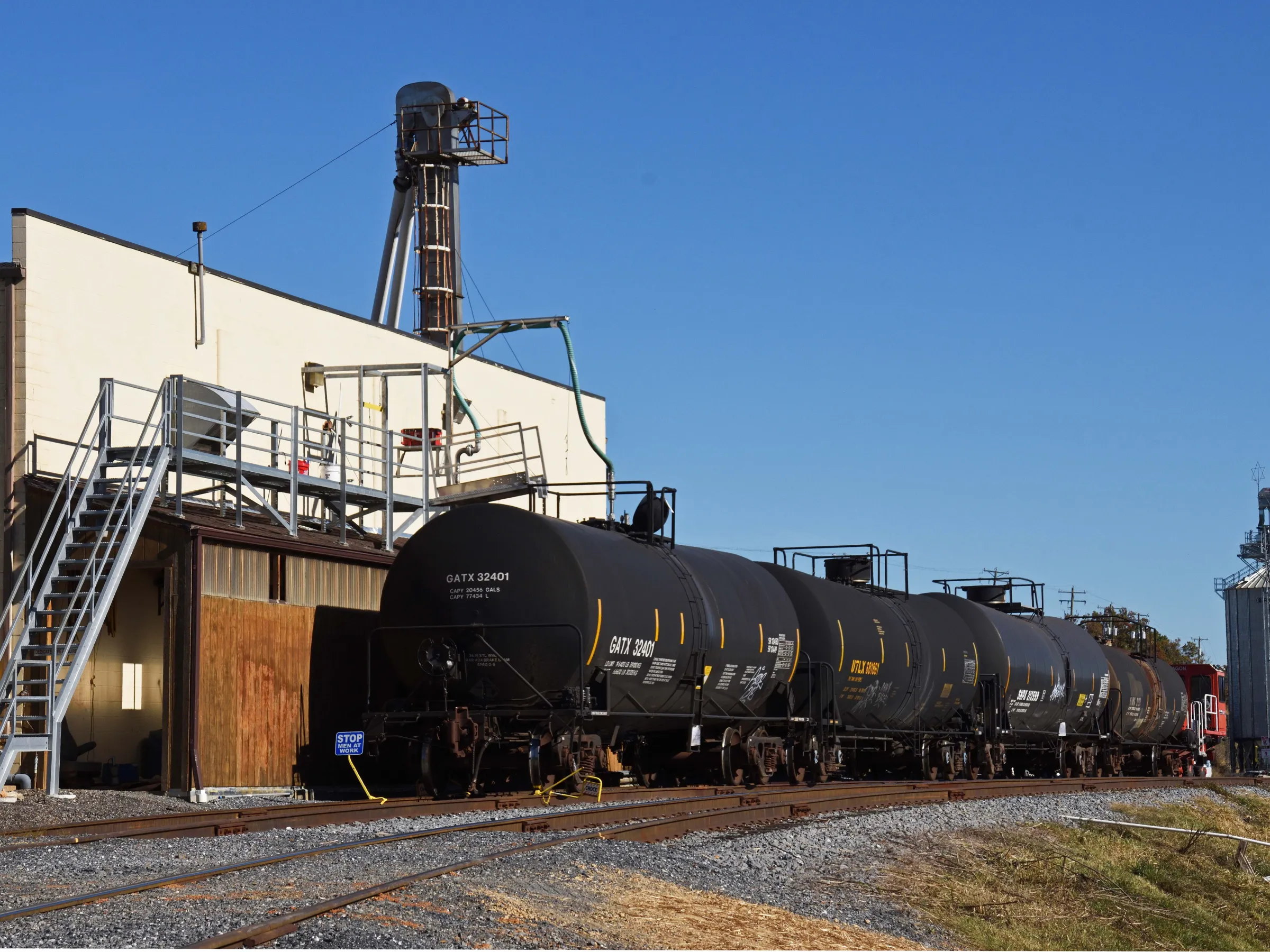 Freight train with black tanker cars next to an industrial building under a clear blue sky.