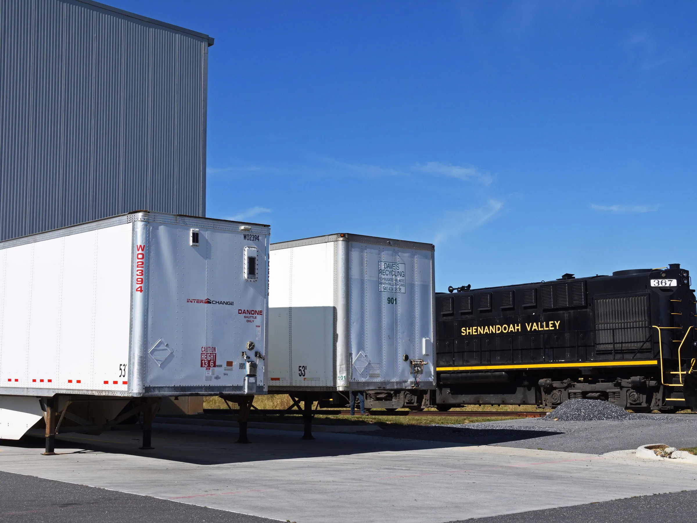 Tractor trailers parked near a black locomotive labeled Shenandoah Valley under a clear blue sky.