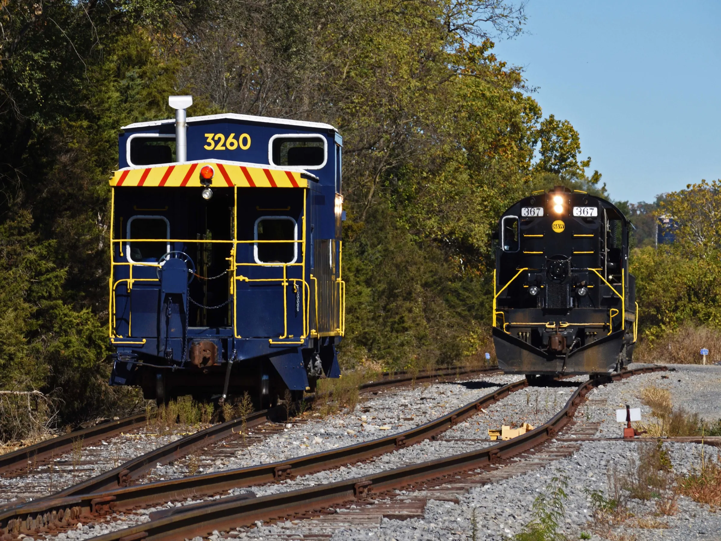 Two trains on separate tracks, one blue caboose with yellow stripes, one black locomotive, surrounded by trees.
