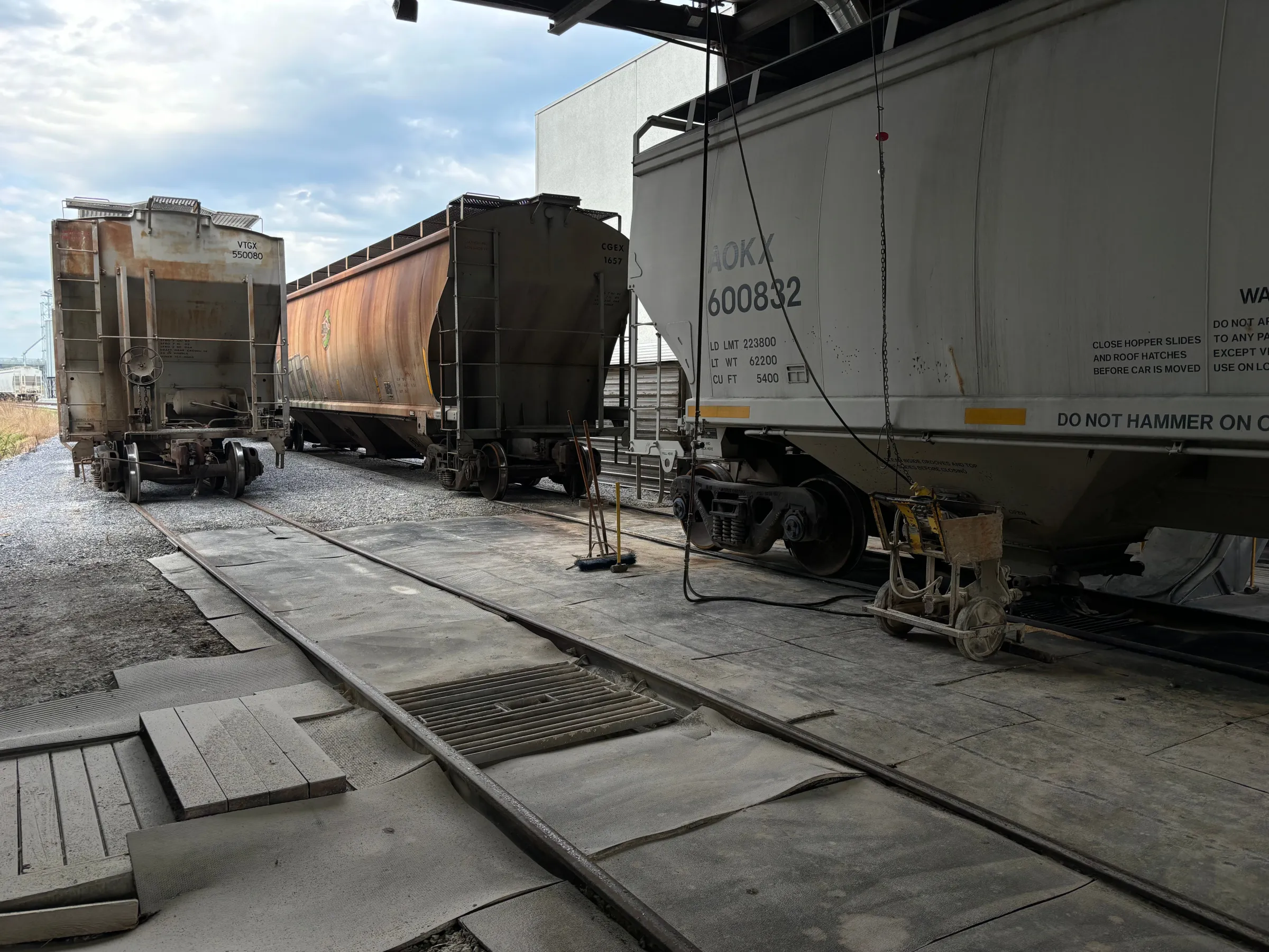 Three freight train cars on tracks under a metal structure with cloudy sky above.