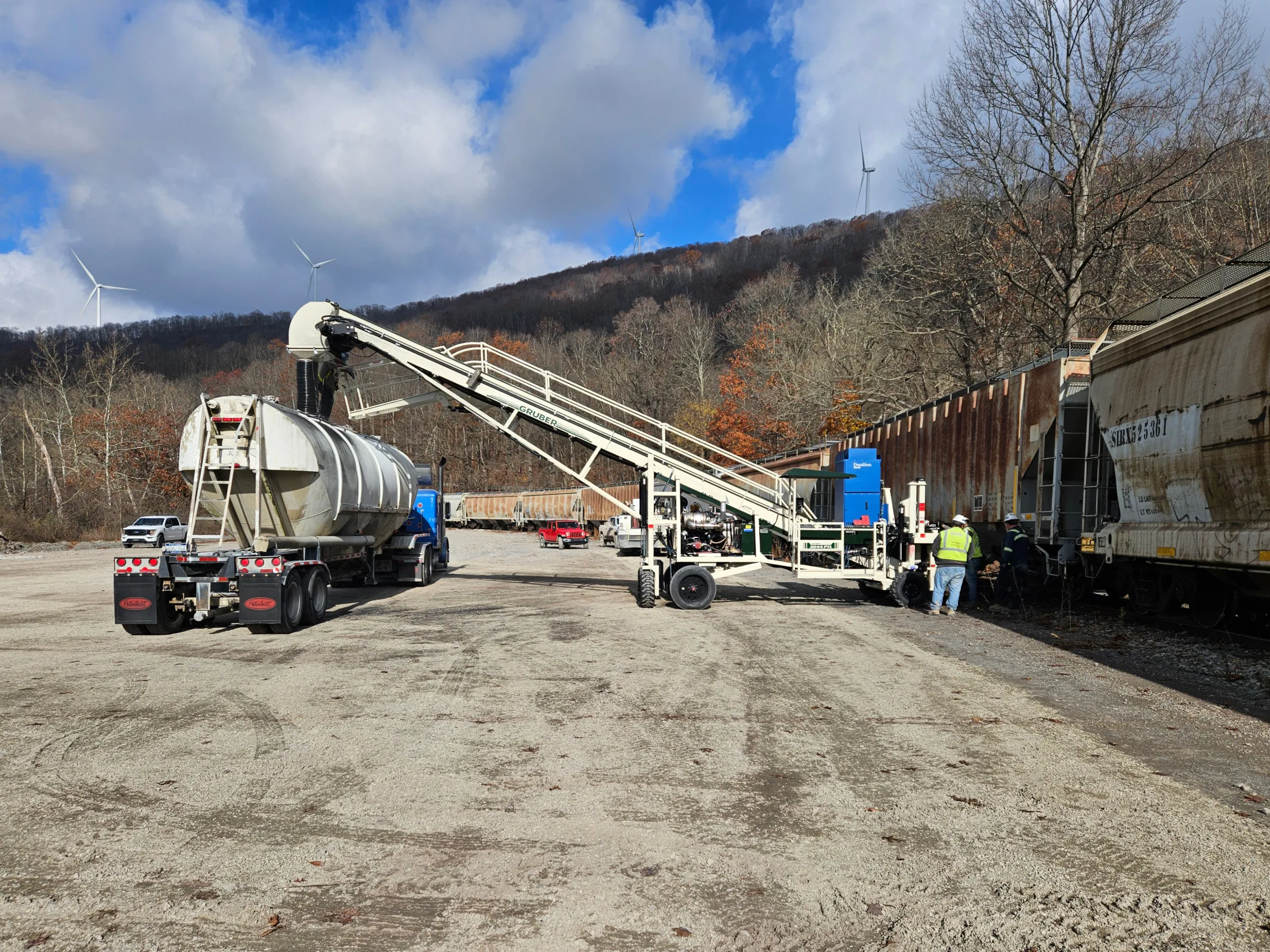 Truck and conveyor transferring materials to rail cars with wind turbines and mountains in the background.