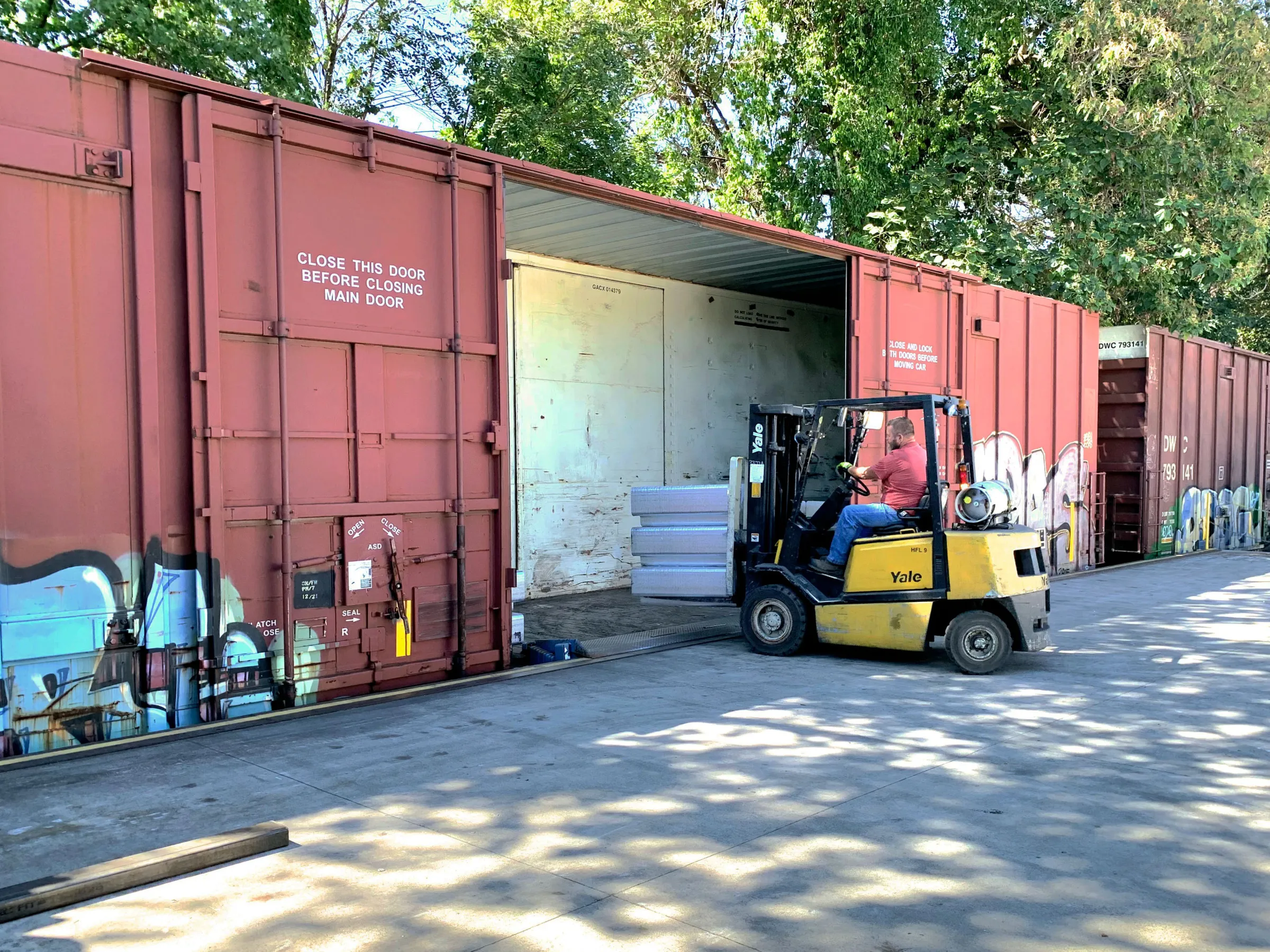 Forklift loading white materials into an open red freight container under trees.