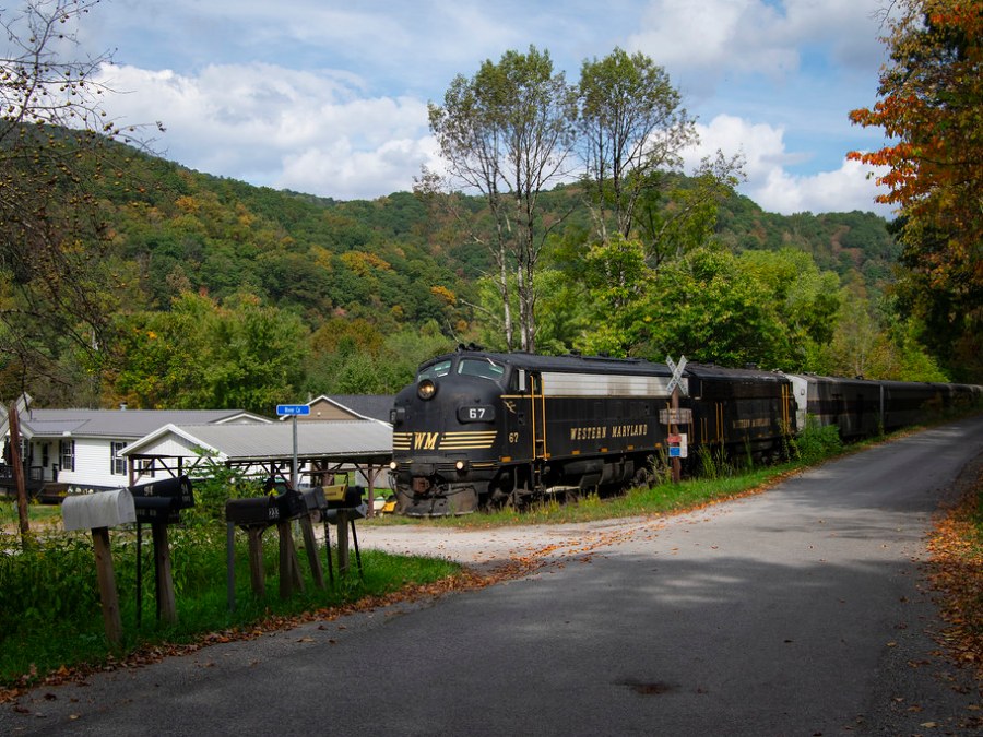Vintage black train with 'Western Maryland' logo passing near houses and mailboxes, surrounded by trees and hills.