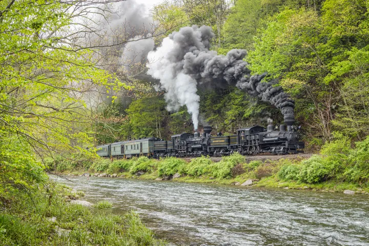Two steam locomotives emit smoke while traveling beside a river in a lush green forest.