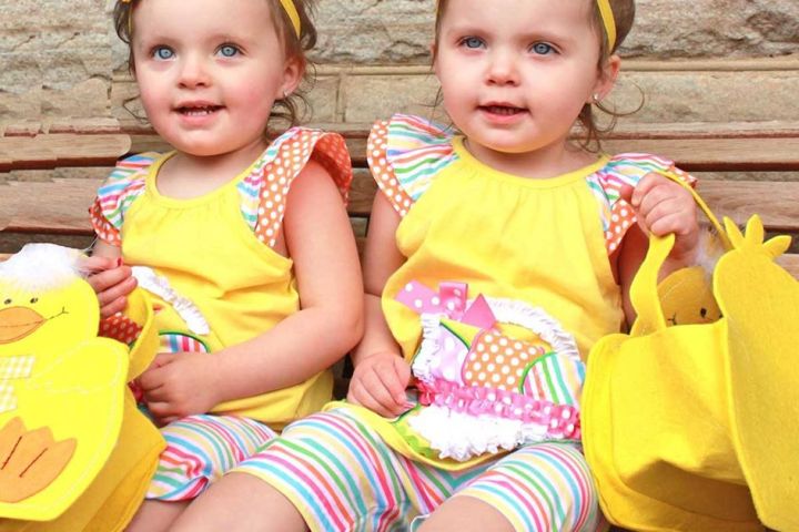 Two toddlers in yellow outfits and flower headbands sitting on a bench holding yellow bags.