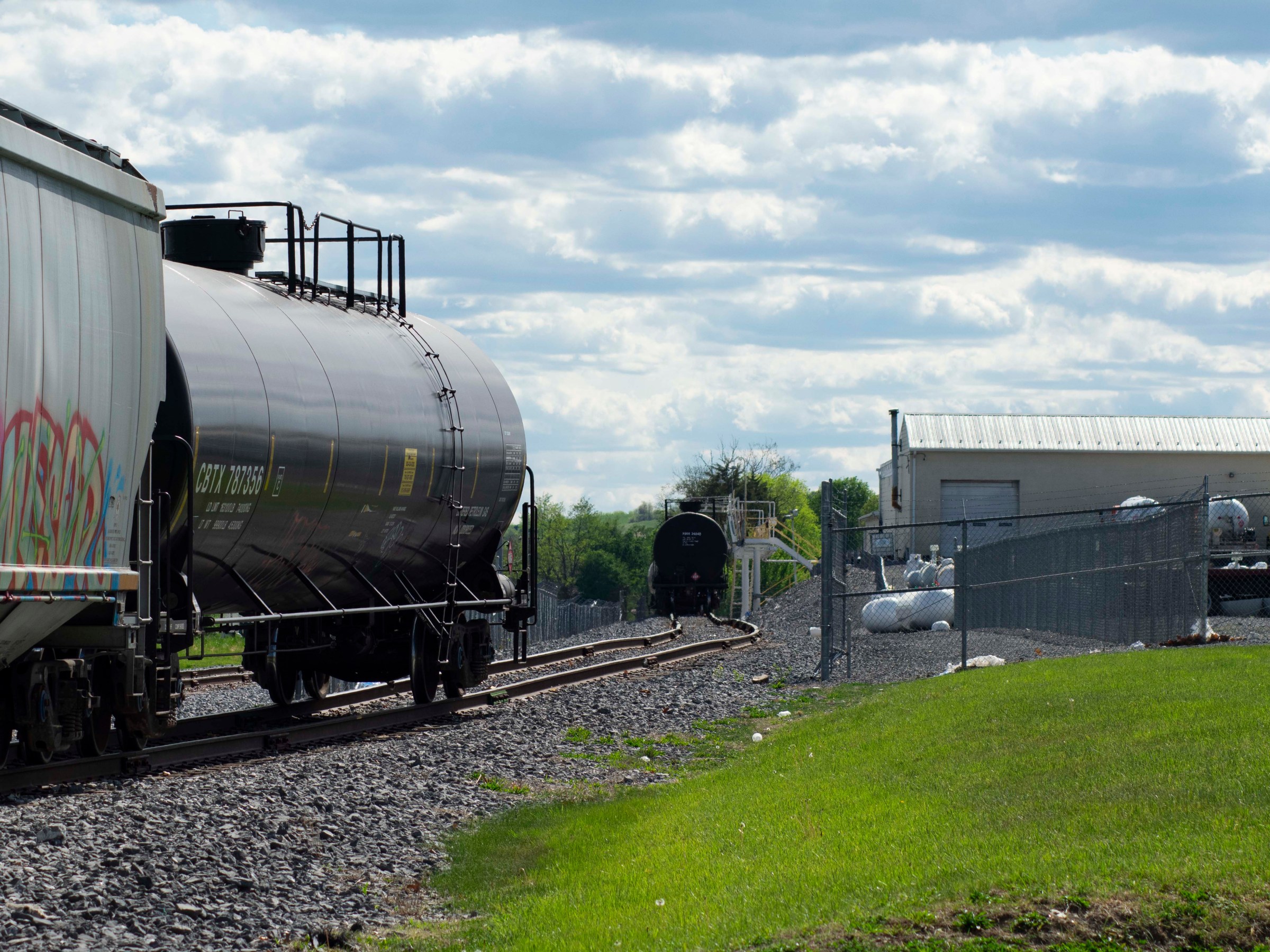 Freight train with tank cars on tracks near a grassy field and industrial building.