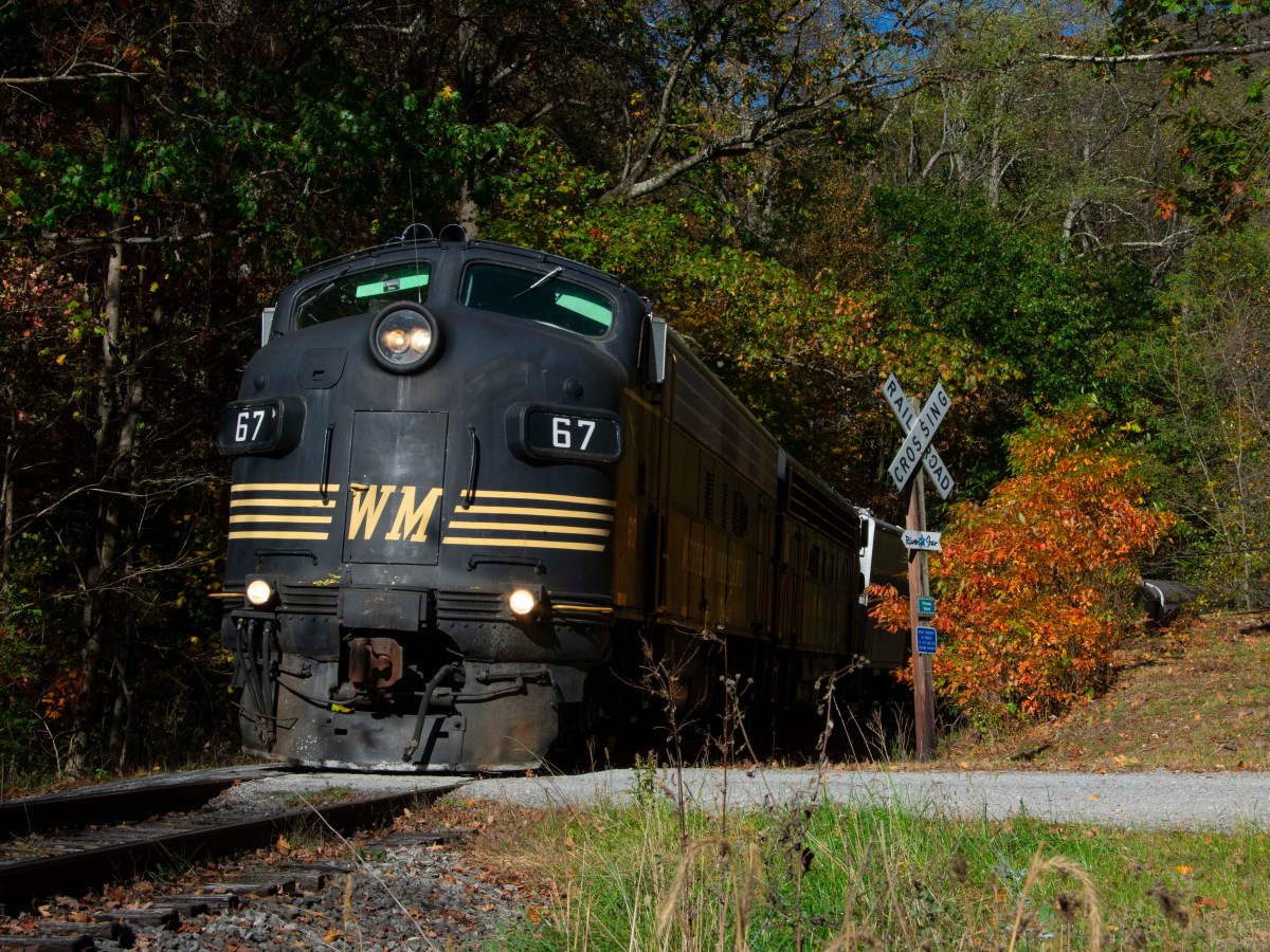 Vintage train with 'WM' logo on tracks through wooded area with autumn trees.
